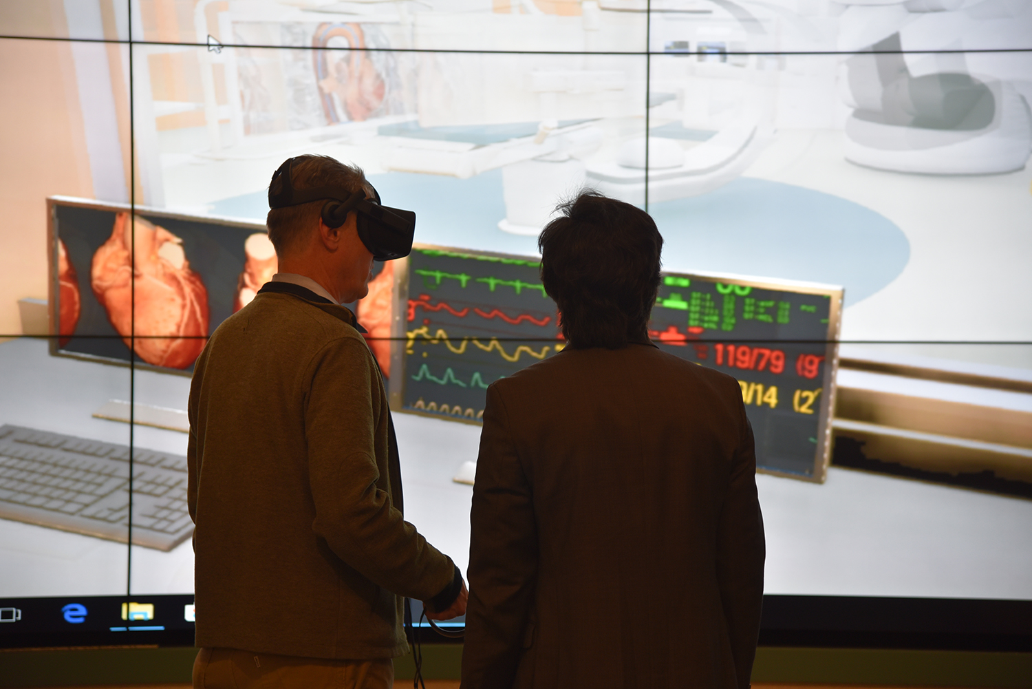 Two men with one wearing virtual reality goggles and screen in background showing design of operating room and EKG monitor