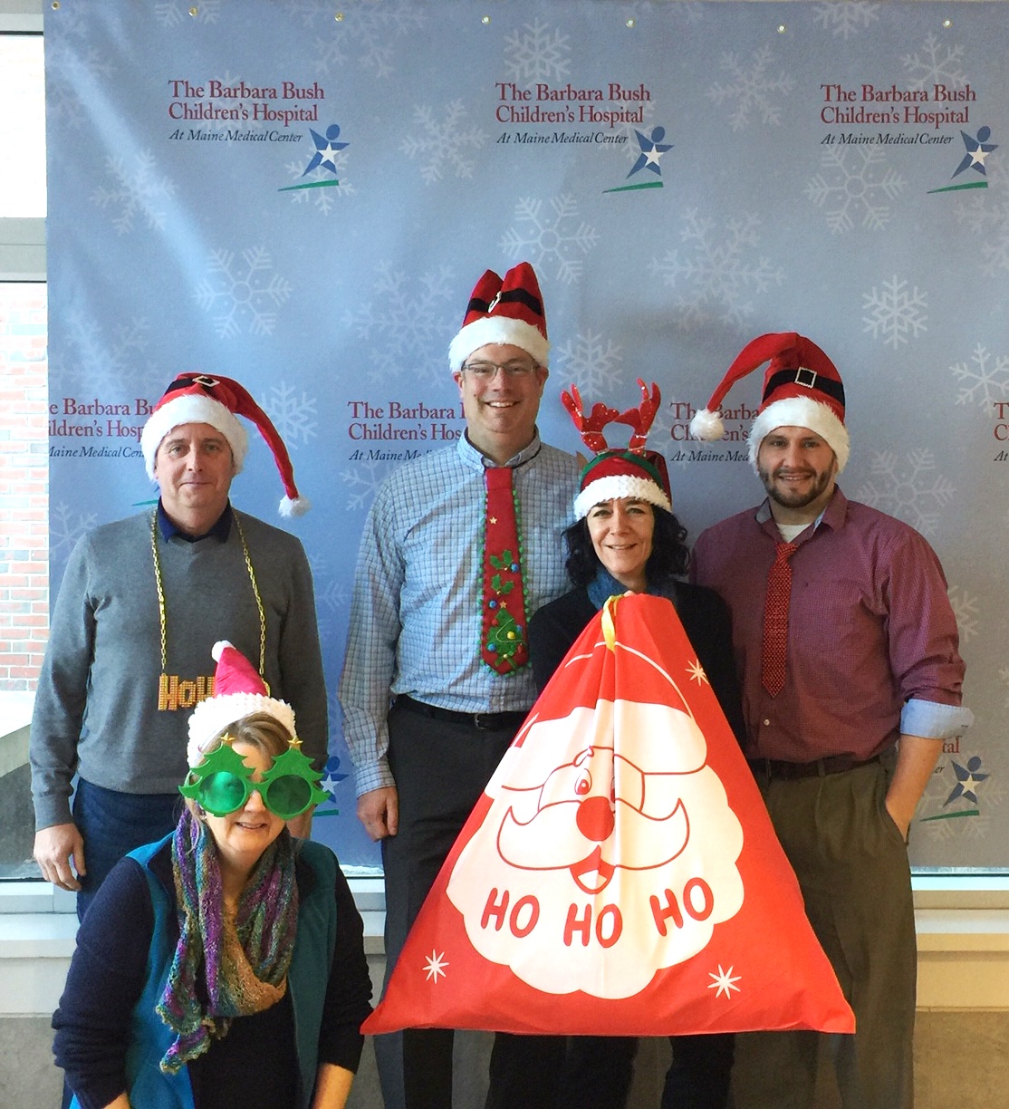 Five e4h team members wearing Christmas hats and holding large red gift bag filled with donations