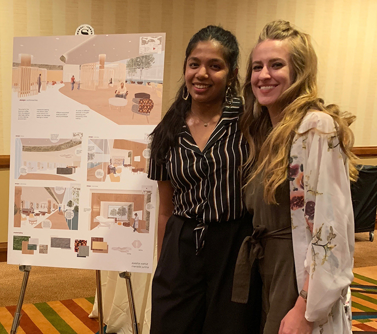Two women smile in front of a poster with a design they created