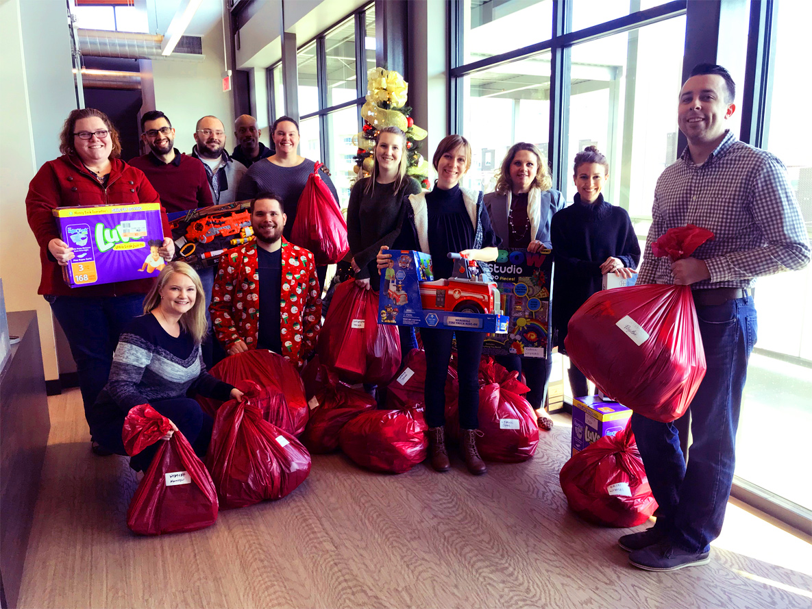 Group of e4h team members in hallway next to windows holding Christmas gifts in red bags