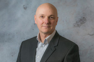 Man smiles while wearing a grey button-down shirt and black blazer - Thomas Morris