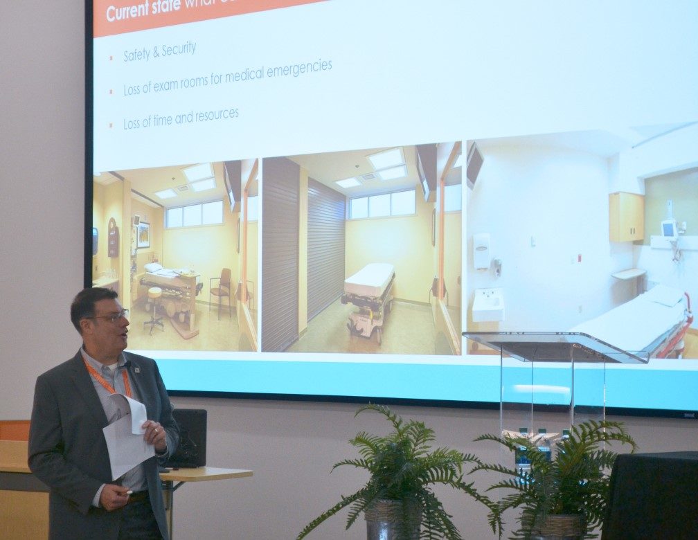 Jon Boyd wears business attire and presents at a conference while looking at large video screen